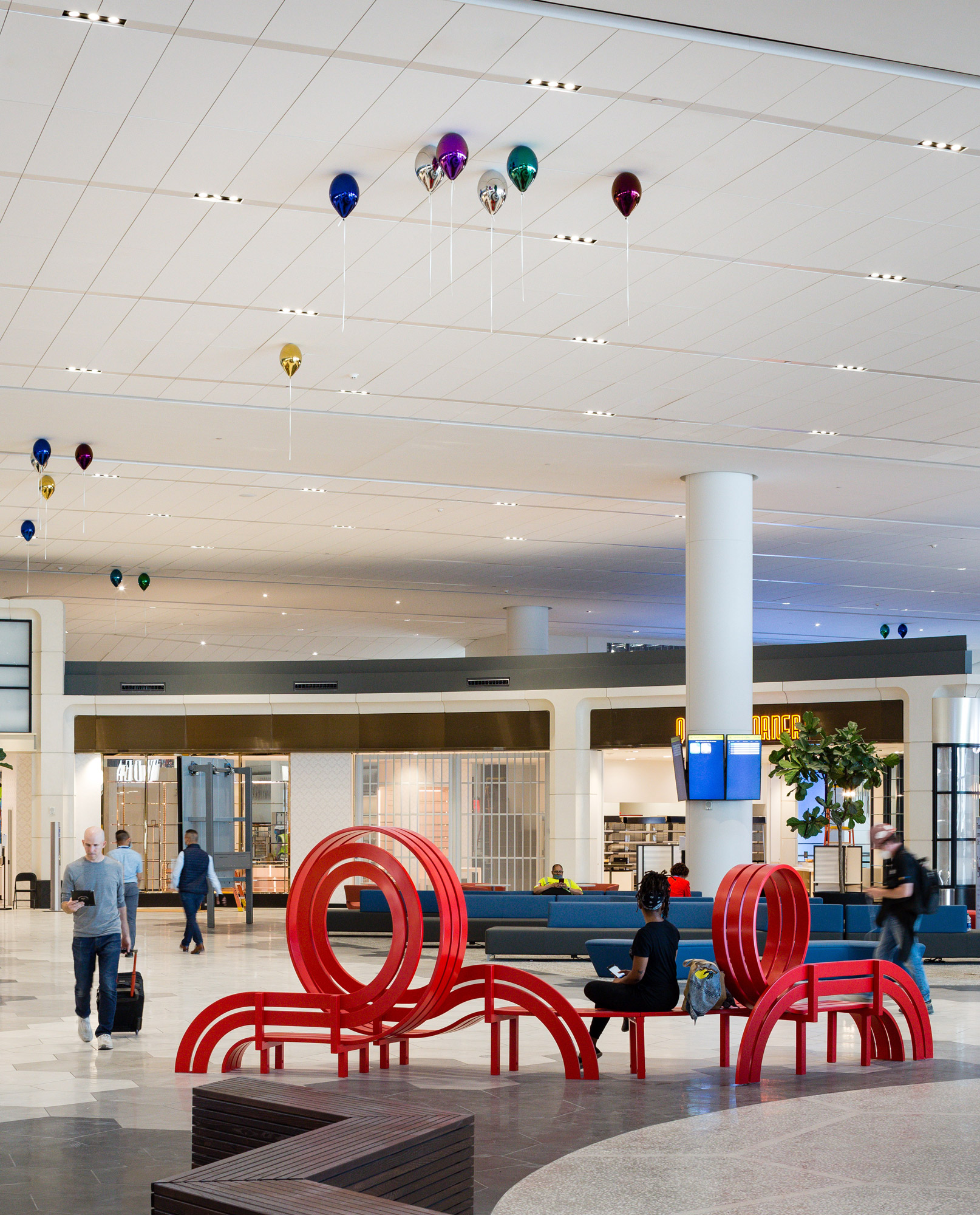 A red sculptural bench and set of metallic colorful balloons pictured in an airport terminal with people milling about.