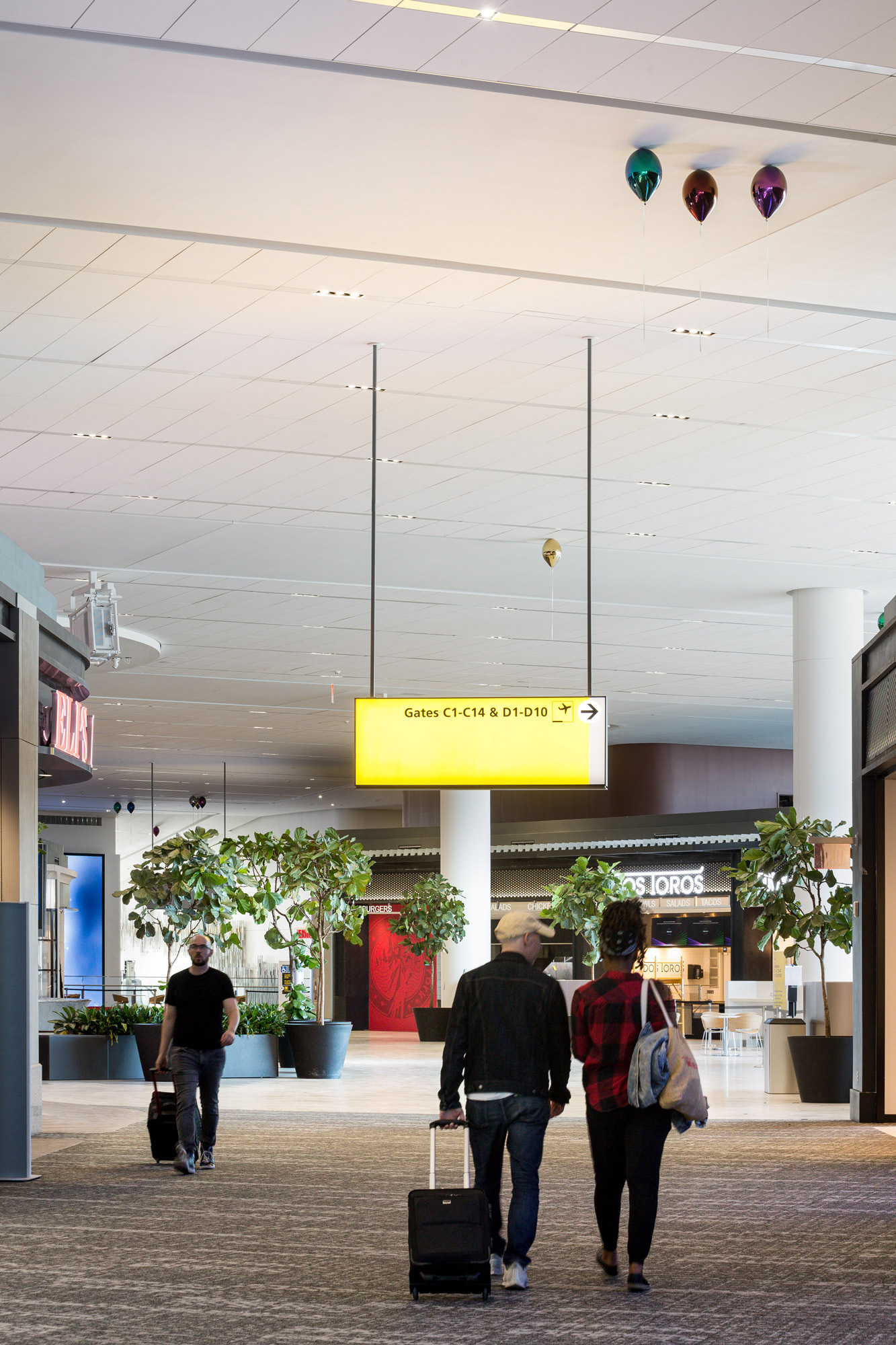 People walk through an airport terminal, underneath tall white ceilings where a series of colorful metallic balloons appear to float.