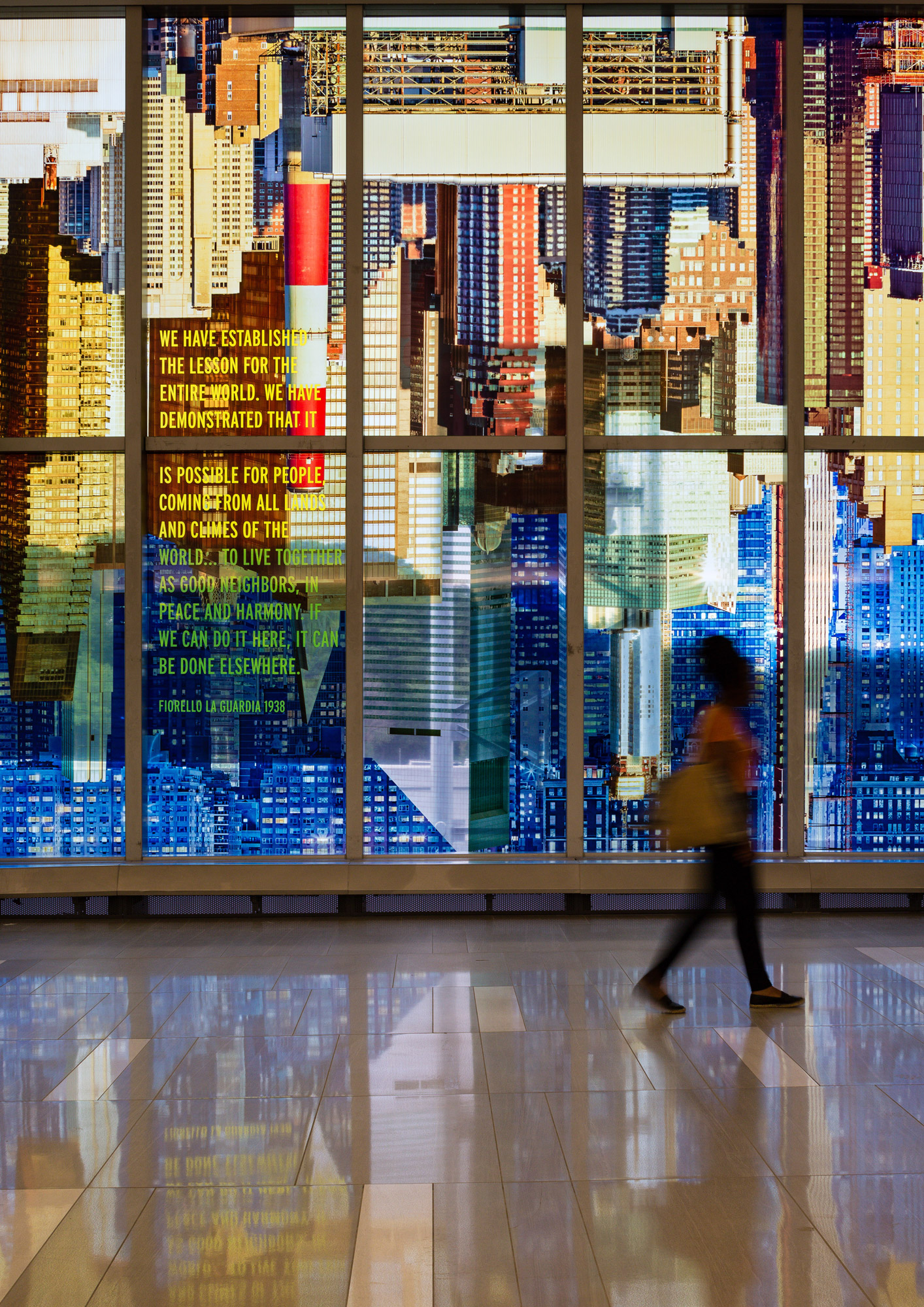 A kaleidoscopic wash of a colorful photo-collaged cityscape fills the length of two stories of tall glass windows in an airport terminal interior.