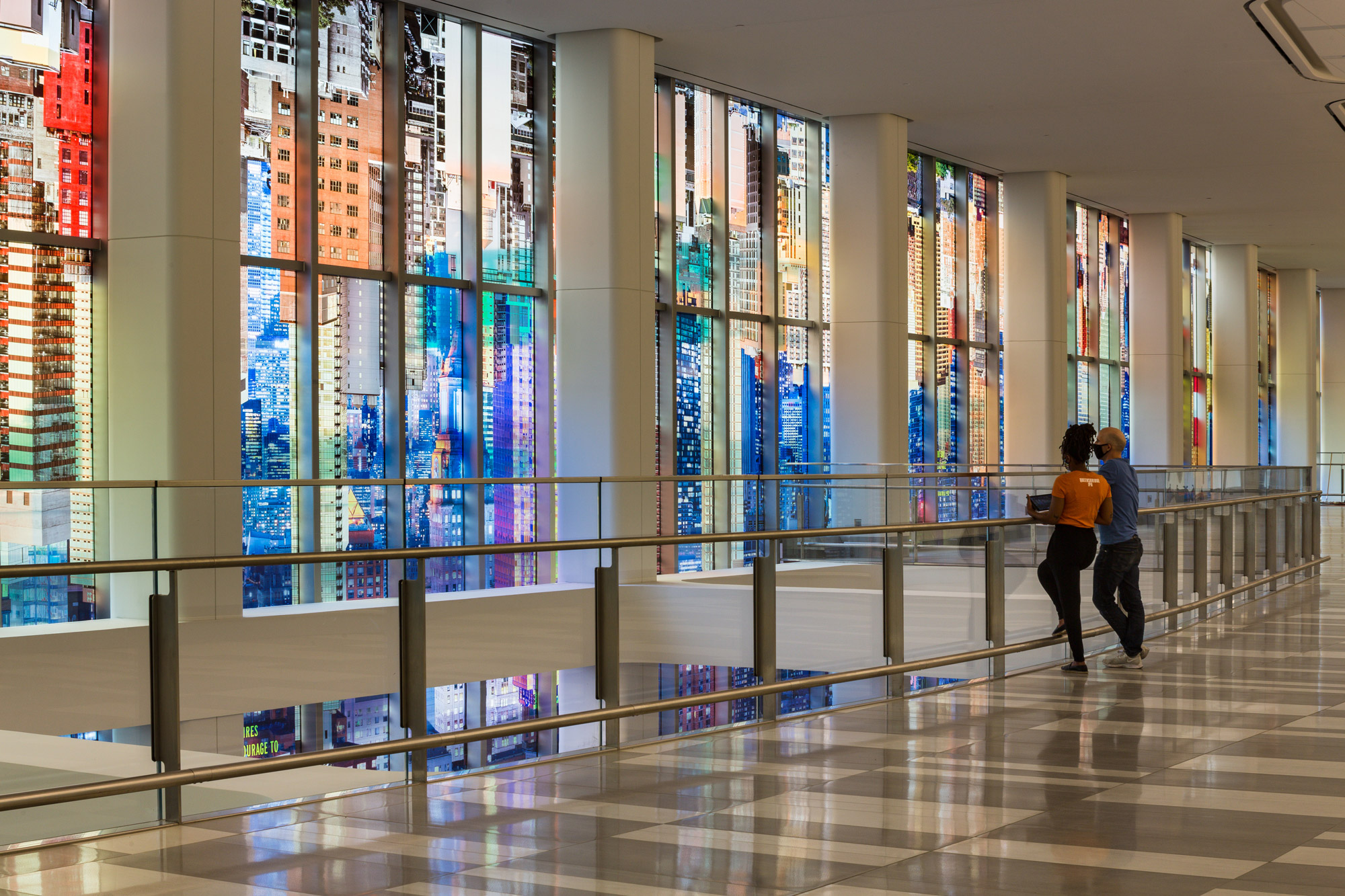 A kaleidoscopic wash of a colorful photo-collaged cityscape fills the length of two stories of tall glass windows in an airport terminal interior.