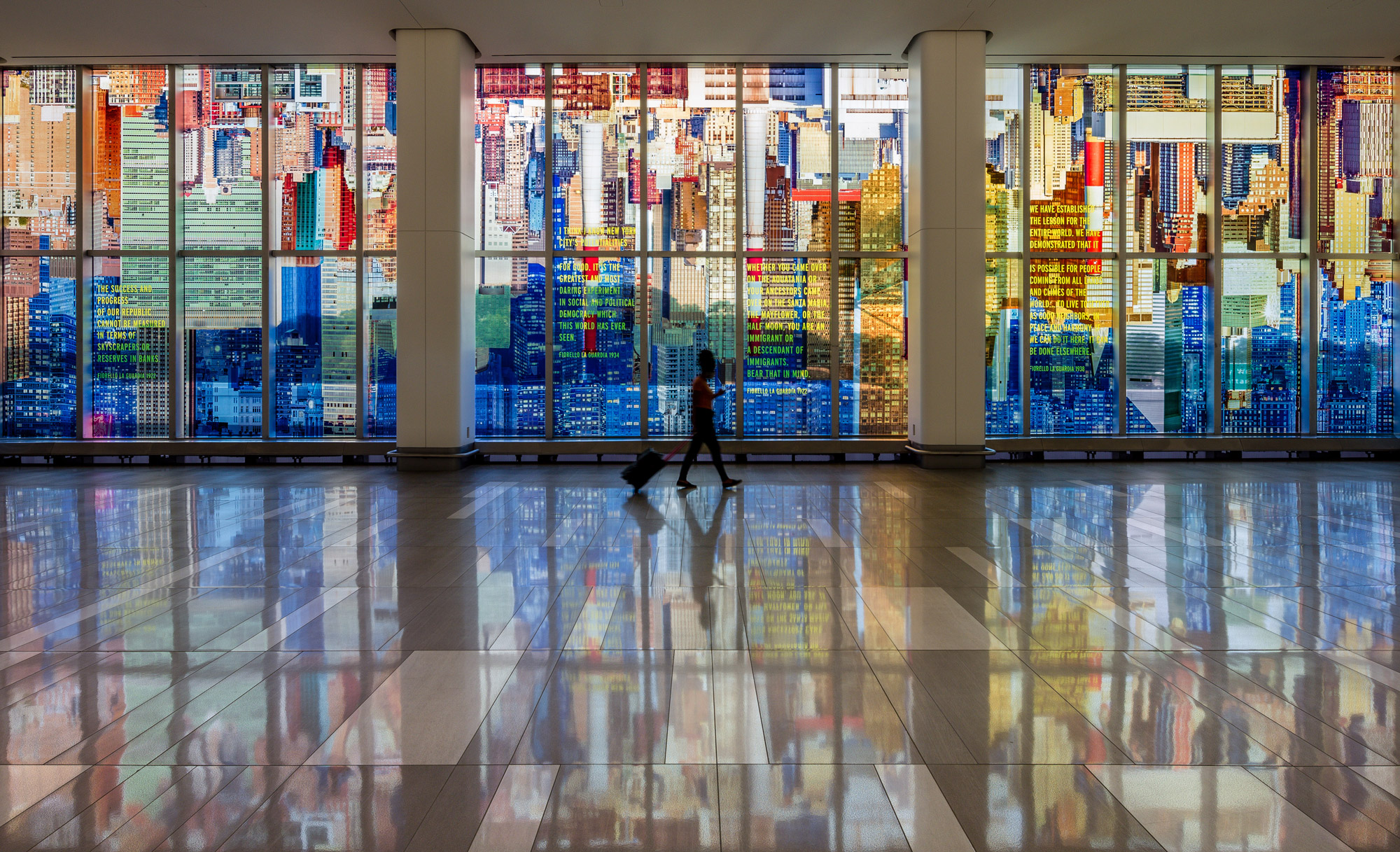 A kaleidoscopic wash of a colorful photo-collaged cityscape fills the length of two stories of tall glass windows in an airport terminal interior.