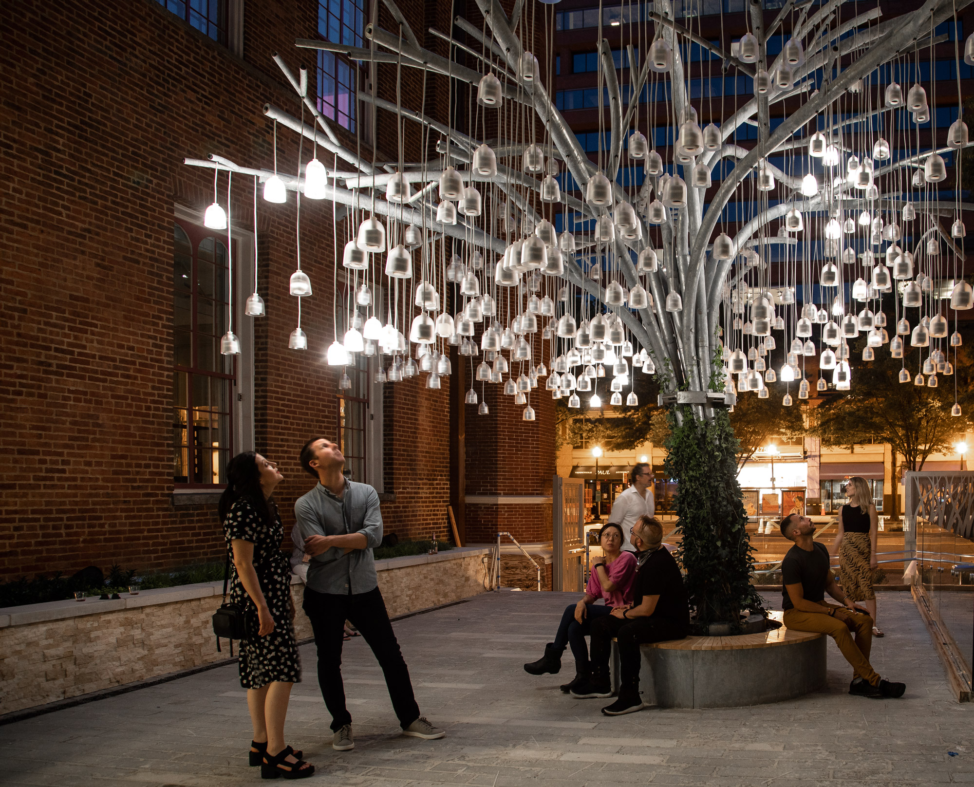 An aluminum sculpture resembling a willow tree with many bell-shaped, lit up speakers hanging from its branches appears to be growing in a courtyard. It is night...