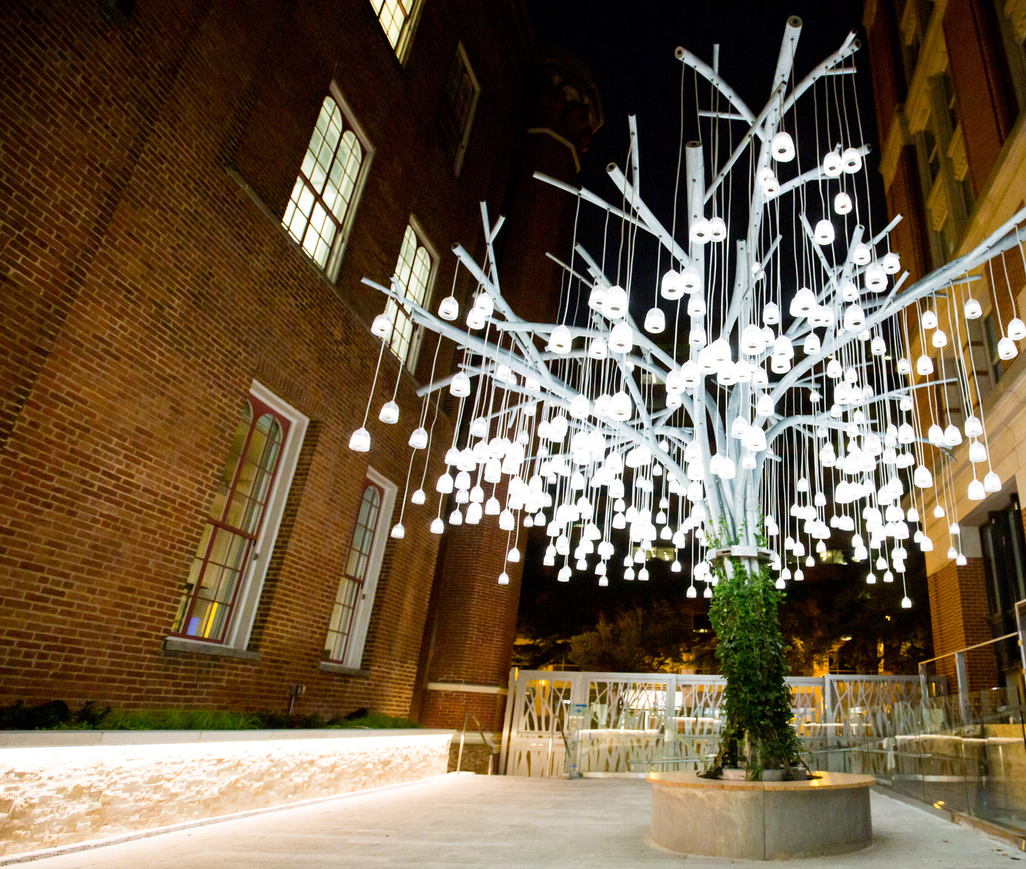 An aluminum sculpture resembling a willow tree with many bell-shaped, lit up speakers hanging from its branches appears to be growing in a courtyard. It is night...
