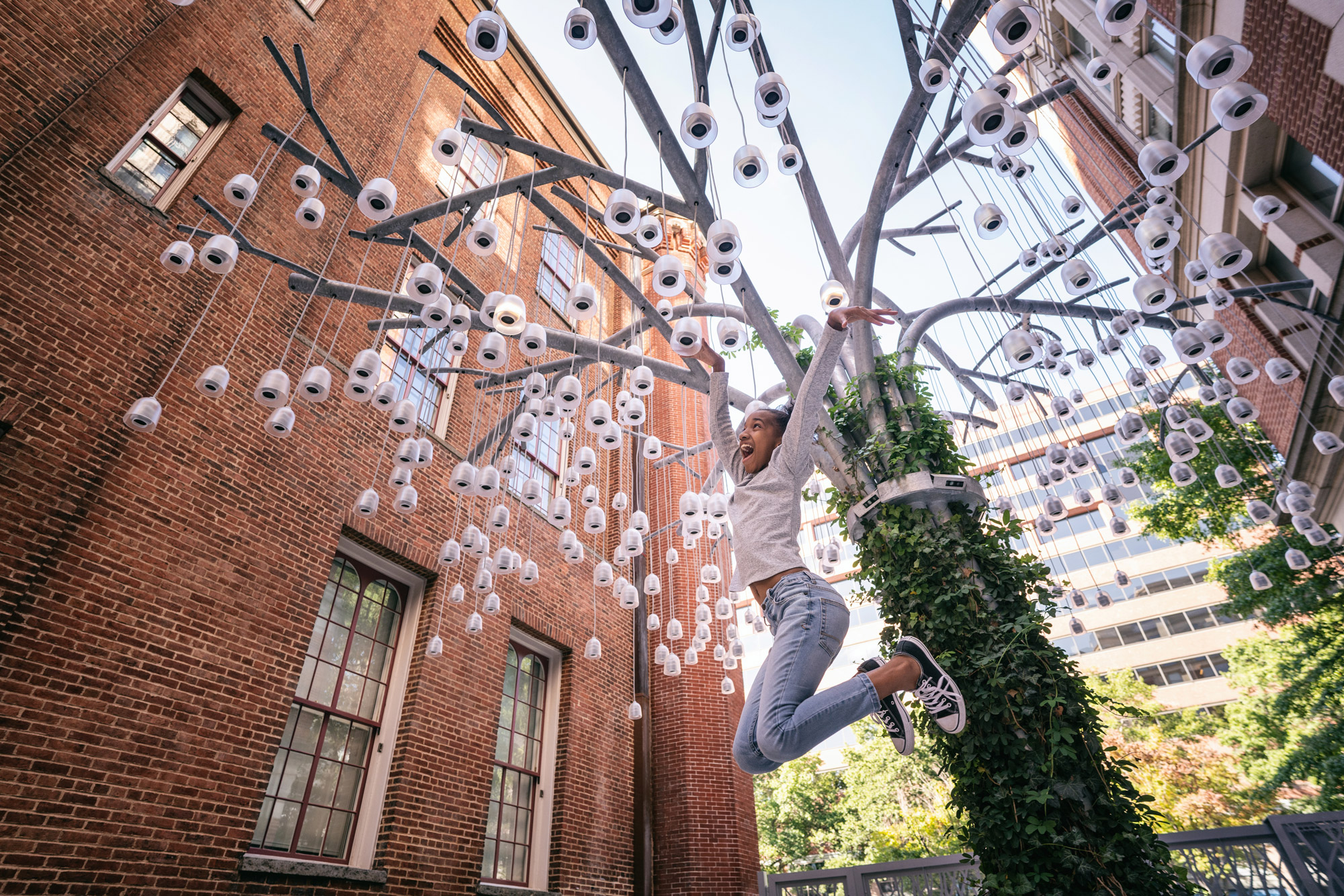 A person is mid-jump, with arms outstretched, in front of an aluminum sculpture resembling a willow tree with many bell-shaped, lit up speakers hanging from its...