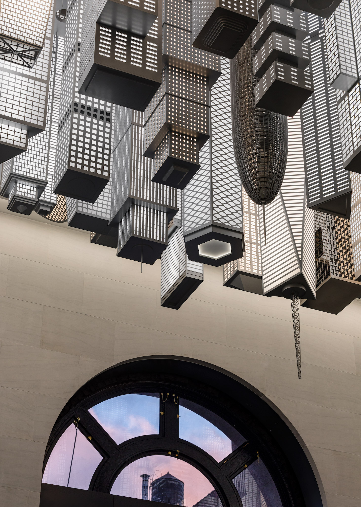 A crop of black and white lit up high-rise buildings descending from the ceiling of the interior of a train station.