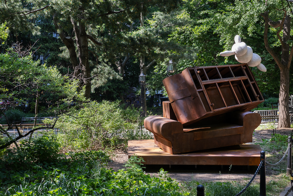 A round rabbit-resembling white form balances atop a steel structure resembling stacked furniture in a green park.