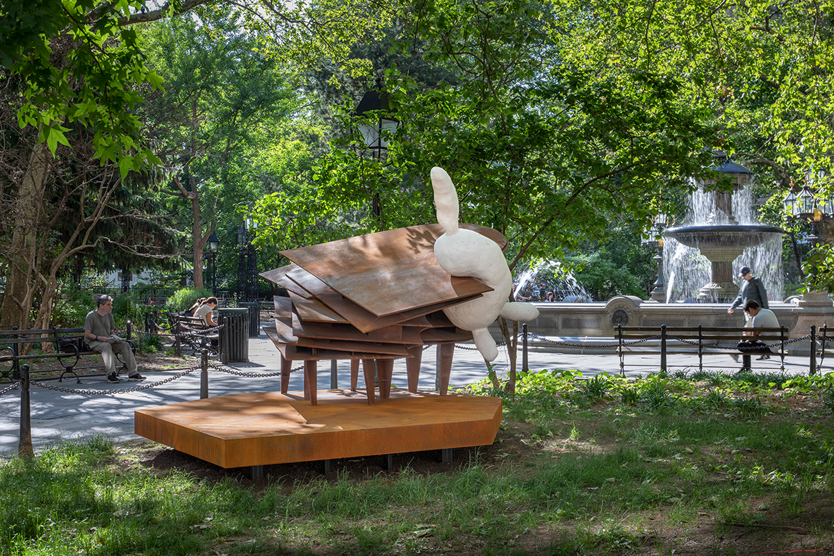 A round rabbit-resembling white form balances atop a steel piano structure in a park with green foliage and a spouting fountain in the background.