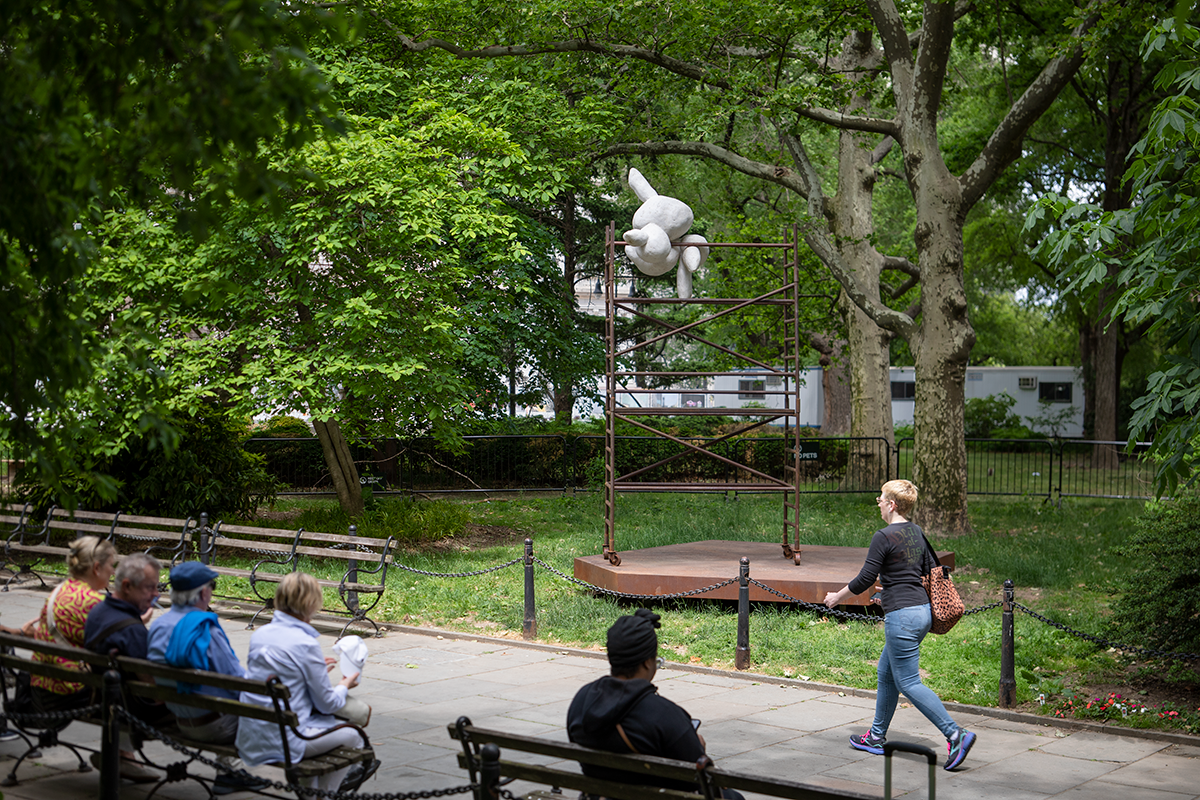 A round rabbit-resembling white form balances atop a steel scaffolding structure in a park, with green foliage in the background. People sit on a bench in front...