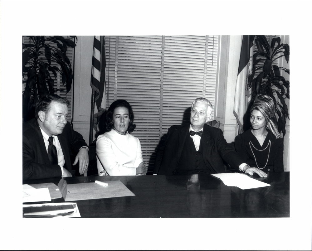 A black and white photograph of two women and two men sitting at a large wood table, in the midst of conversation.