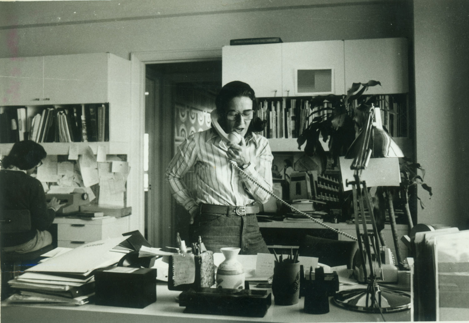 A black and white photo of a woman in business casual attire answering the phone while standing in an office.