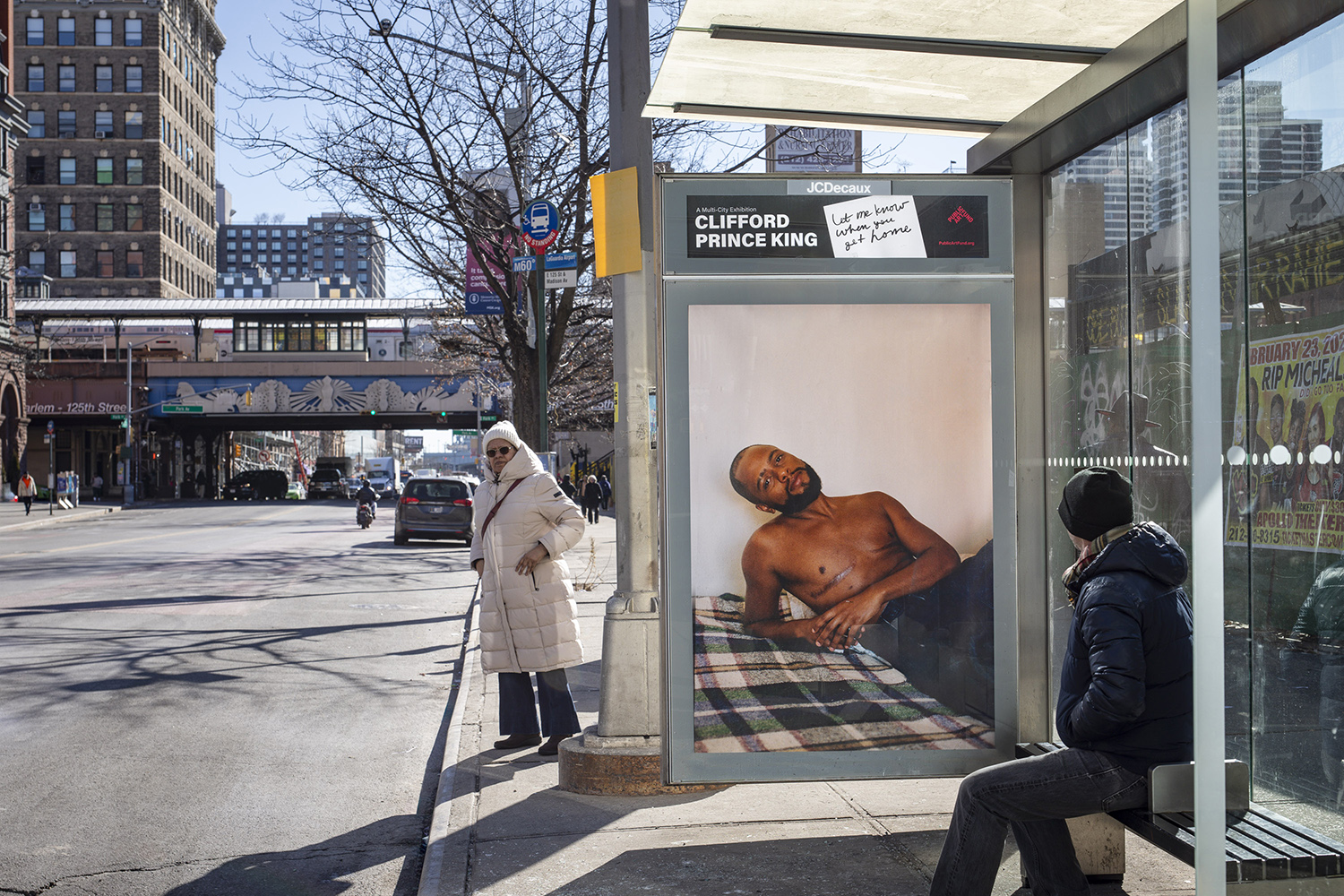 混雑した街のバス停に描かれた写真には、胸筋に傷のある上半身裸の黒人男性が、緑のチェック柄の布が敷かれたベッドにもたれかかり、こちらを見ている...