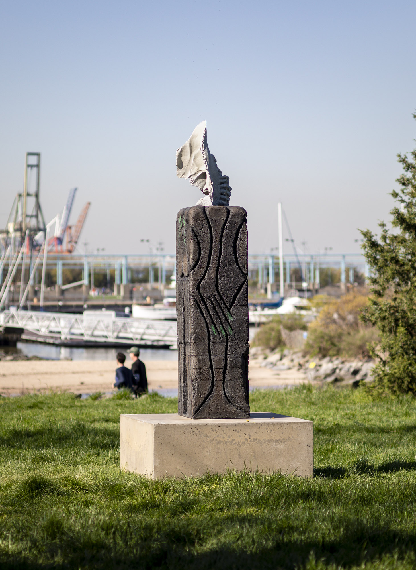 A close-up shot of a patinated bronze sculpture with a square concrete base installed along a grassy terrace. On top of the sculpture sits a recreated off-white...
