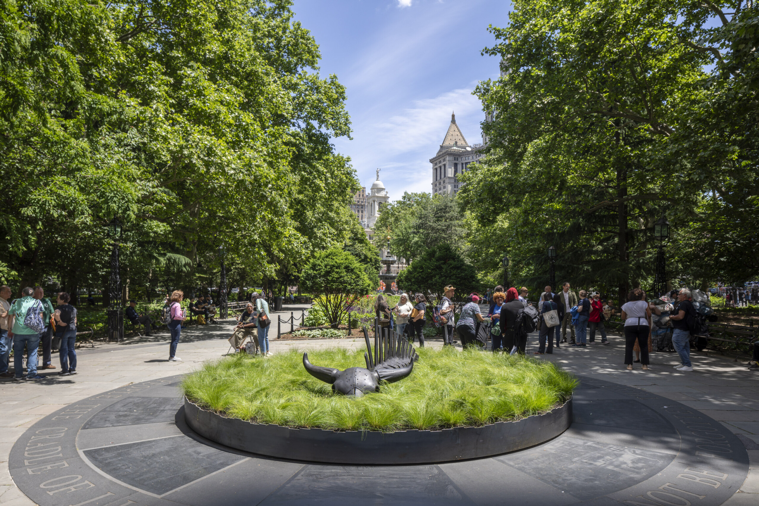 A 10-foot-long black steel sculpture of a bison skeleton sits within a shallow circular bed of green grass in a public park in New York City.