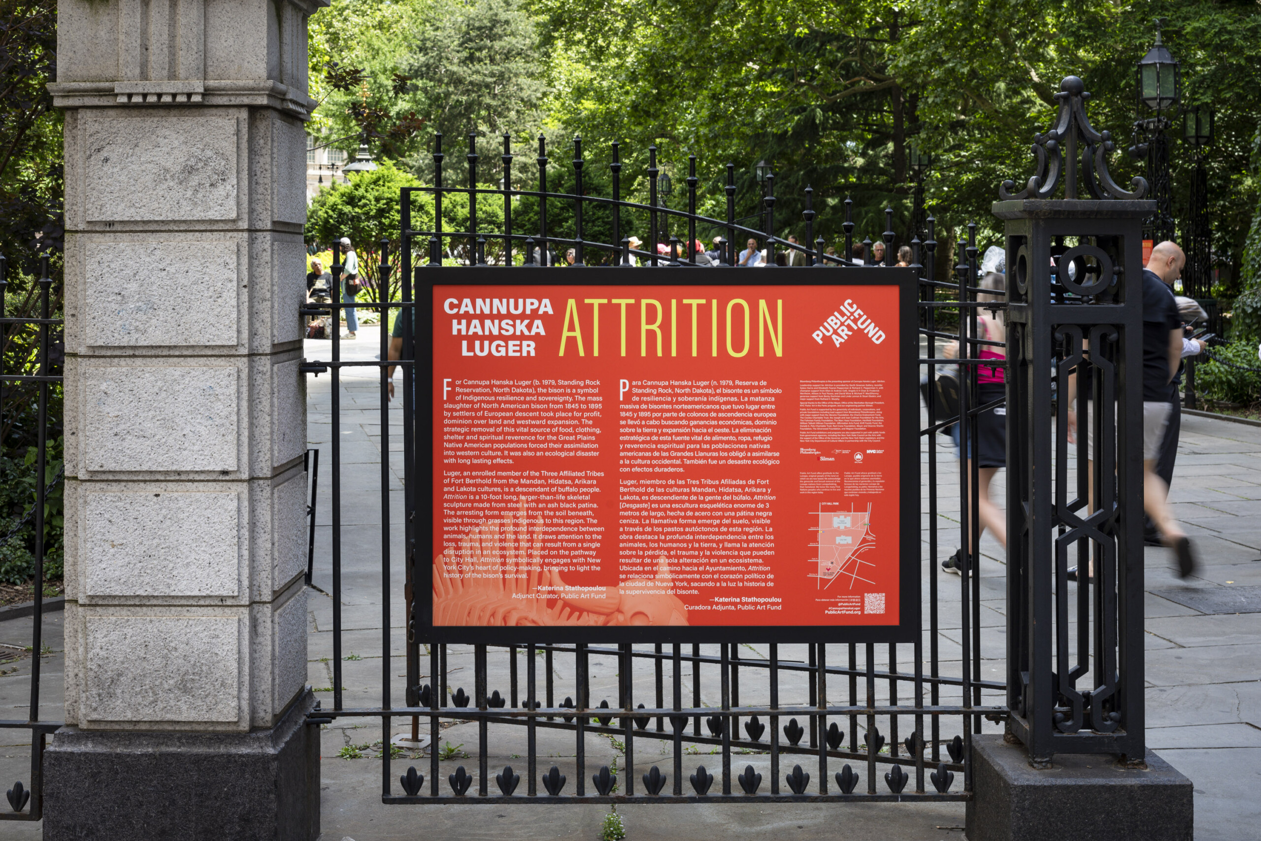 An orange wayfinding sign installed in a black frame and attached to a black, metal gate at a New York City park.
