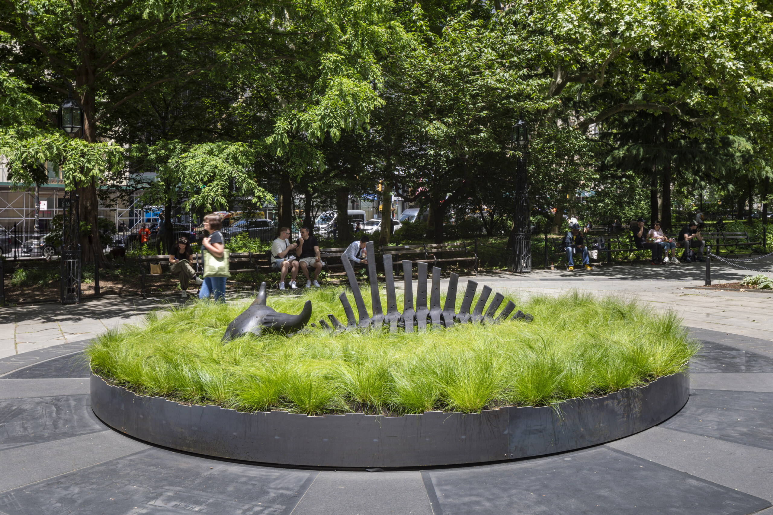 A 10-foot-long black steel sculpture of a bison skeleton sits within a shallow circular bed of green grass in a public park in New York City.